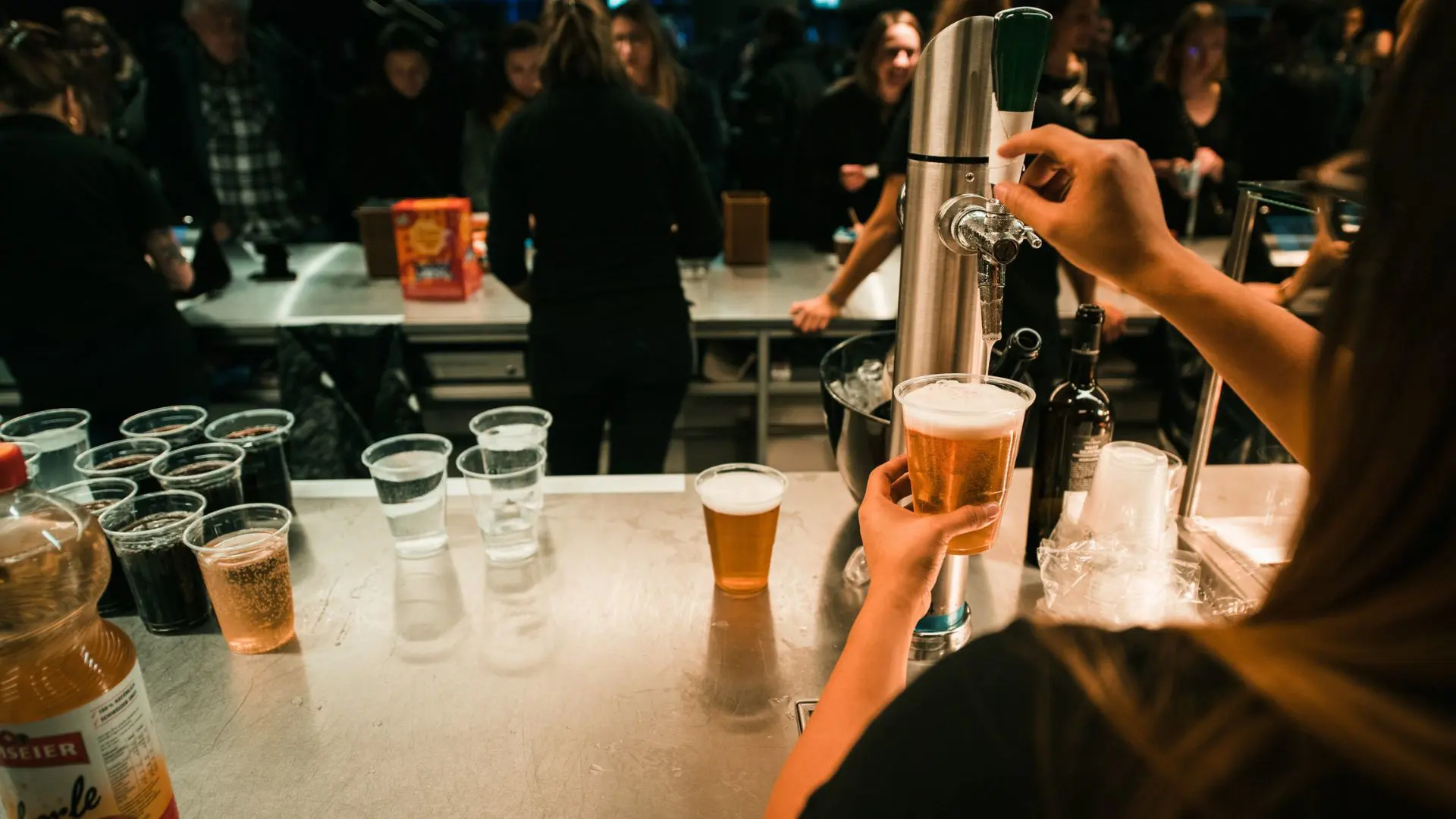 Eine Mitarbeiterin zapft an der Bar ein Bier, während im Hintergrund Gäste auf ihre Getränke warten.